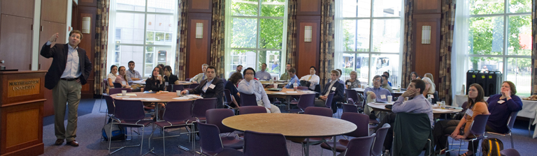 people in seminar sitting at round tables in meeting room watching a speaker present